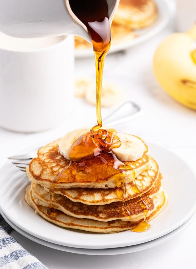 A stack of pancakes topped with bananas and syrup being poured over the top. 