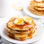 A stack of banana pancakes on a plate topped with banana slices and syrup.