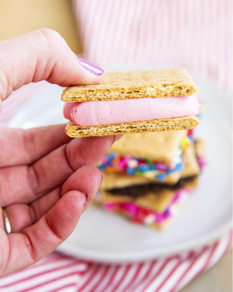 A hand holding a graham cracker cookie sandwich, which is two graham cracker halves full of strawberry frosting in the middle.