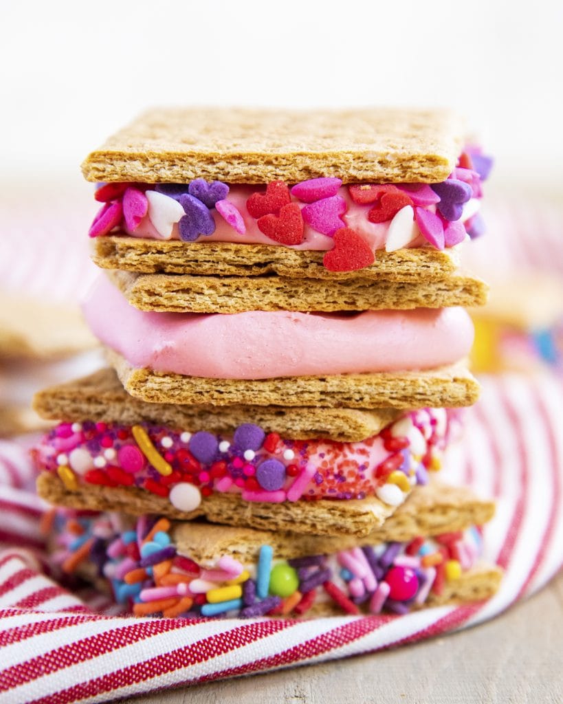 A stack of four graham cracker cookie sandwiches with pink frosting, and pink and purple sprinkles on them.