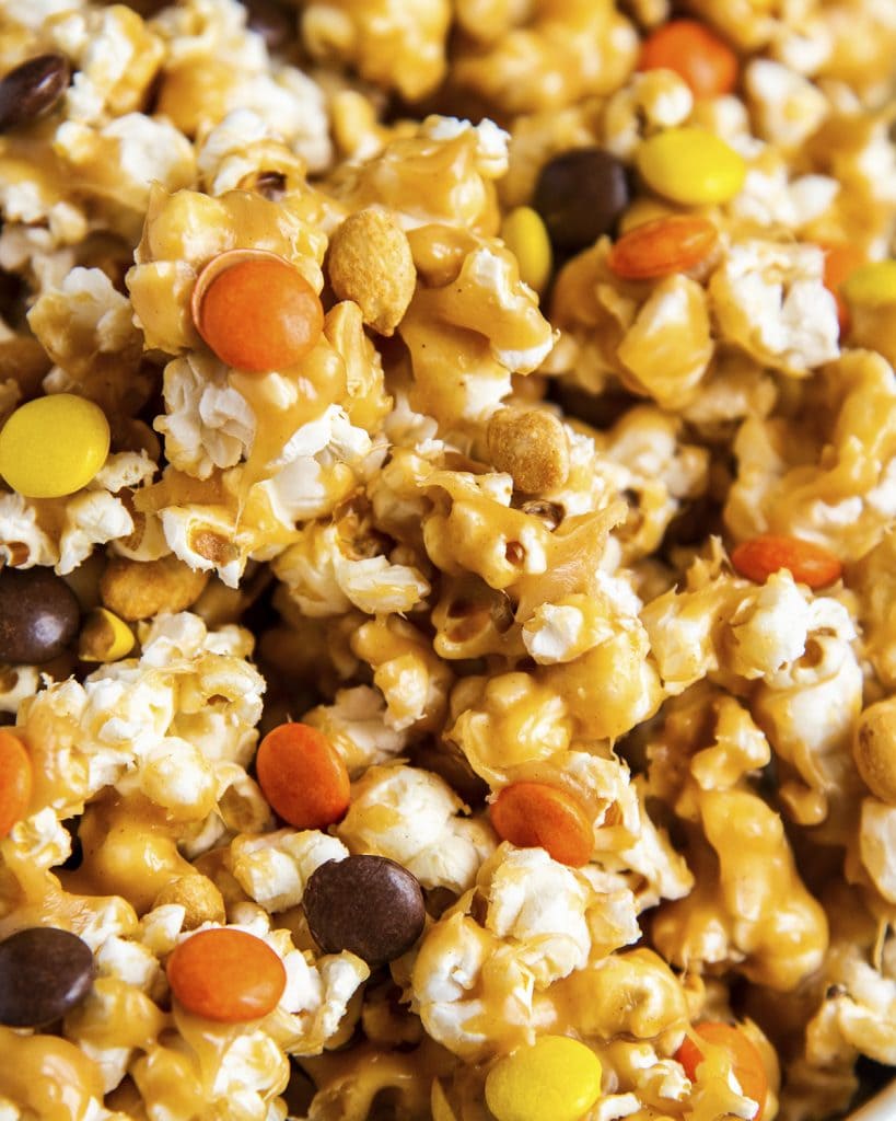 A close up of popcorn covered in an orange brown peanut butter mixture, and full of orange, brown, and yellow reeses pieces candies.