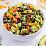 A square image of a bowl of avocado and black bean salad with tomato and pepper pieces.