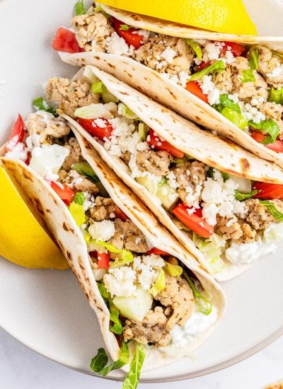 A close up of Greek chicken tacos topped with lettuce, feta, and tomatoes.