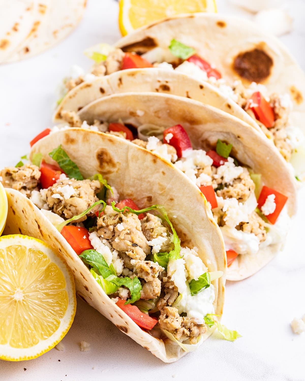 Greek Chicken tacos in tortillas squished next to each other topped with lettuce and feta cheese.