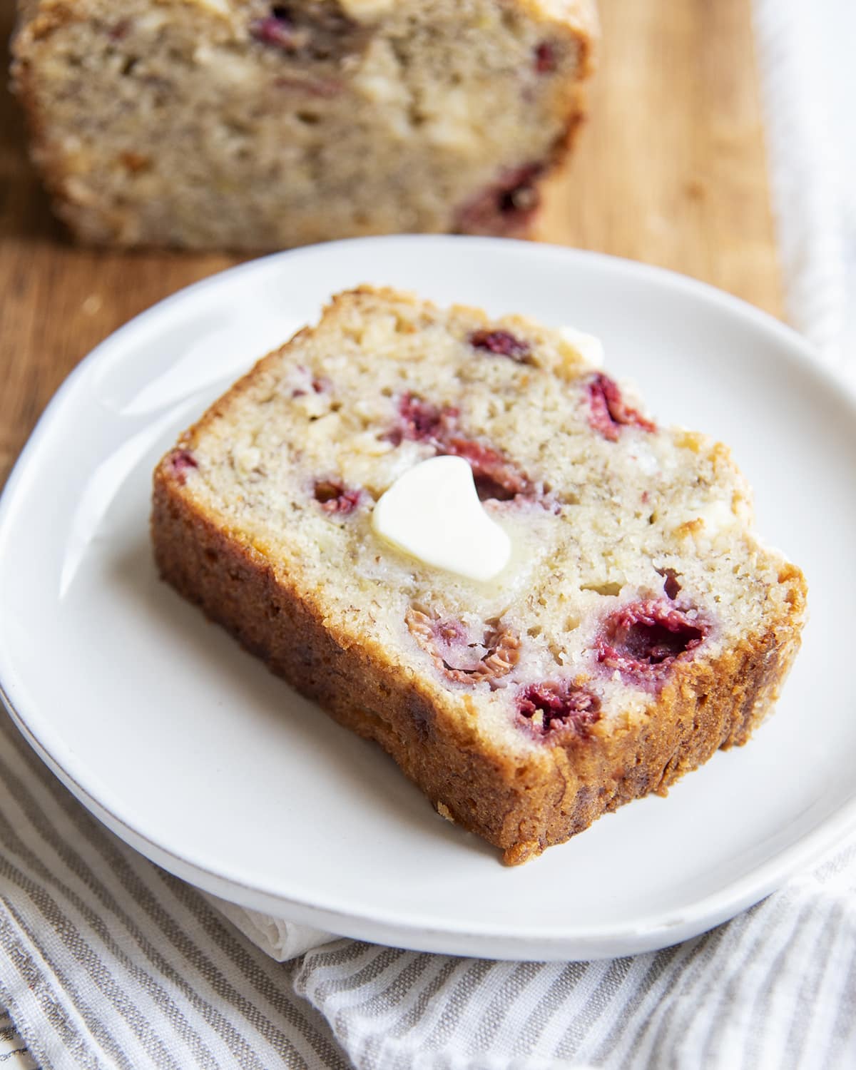 A slice of banana bread with raspberries and white chocolate chips with a melted butter slab on top.