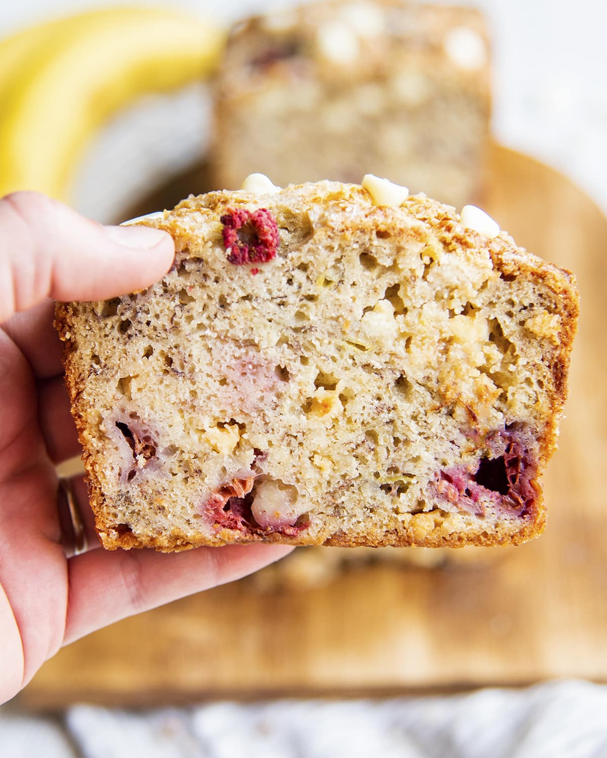 A hand holding a slice of banana bread with pieces of raspberries and white chocolate chips in it.