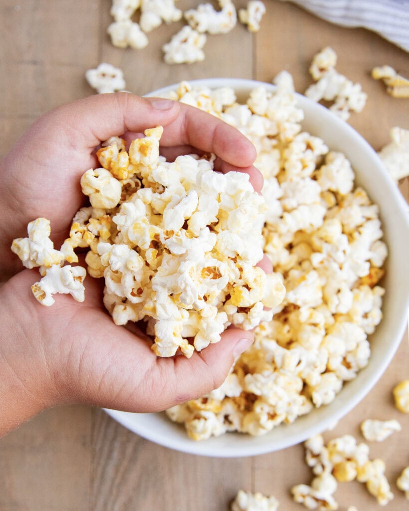 Two hands holding some kettle popcorn in them, above a bowl of it.
