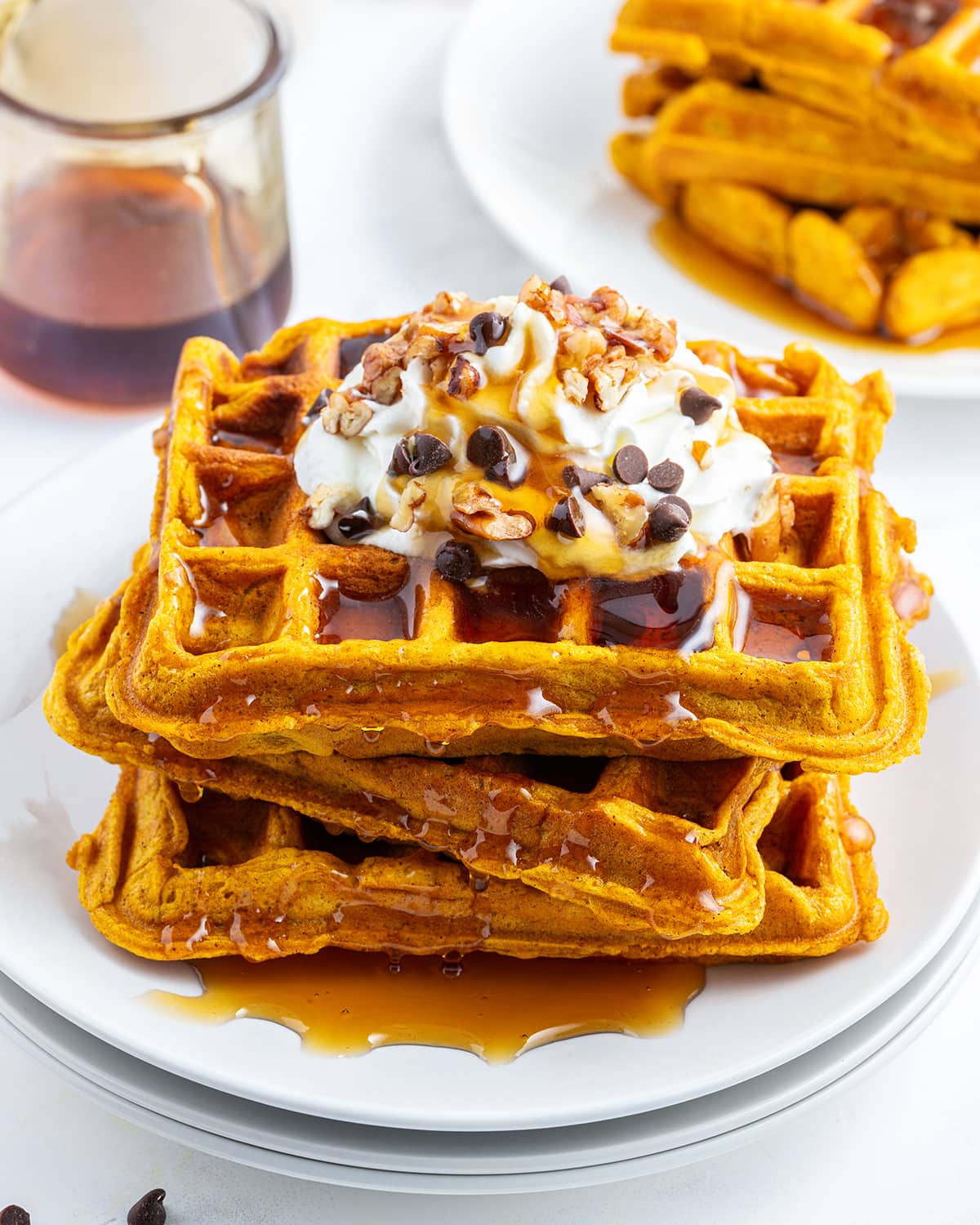A stack of pumpkin waffles on a plate topped with whipped cream, syrup, and mini chocolate chips.