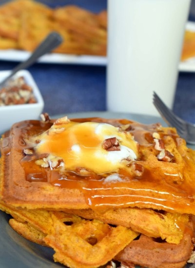 4 pumpkin waffles layered on a blue plate topped with whipped cream, pecans, and syrup with a glass of milk in the background
