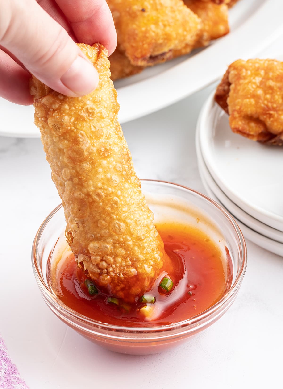 A hand dipping an egg roll into a bowl of sweet and sour sauce.