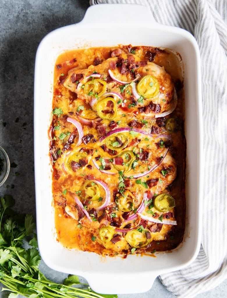 A baking pan of BBQ baked chicken breast topped with cheese, red onion, and jalapenos.