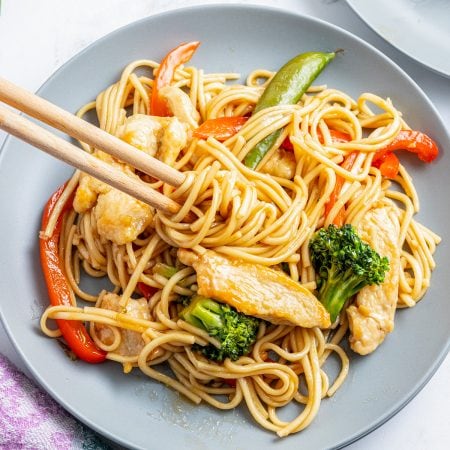 An overhead photo of a plate of chicken lo mein with vegetables with chopsticks wrapping up some of the noodles.