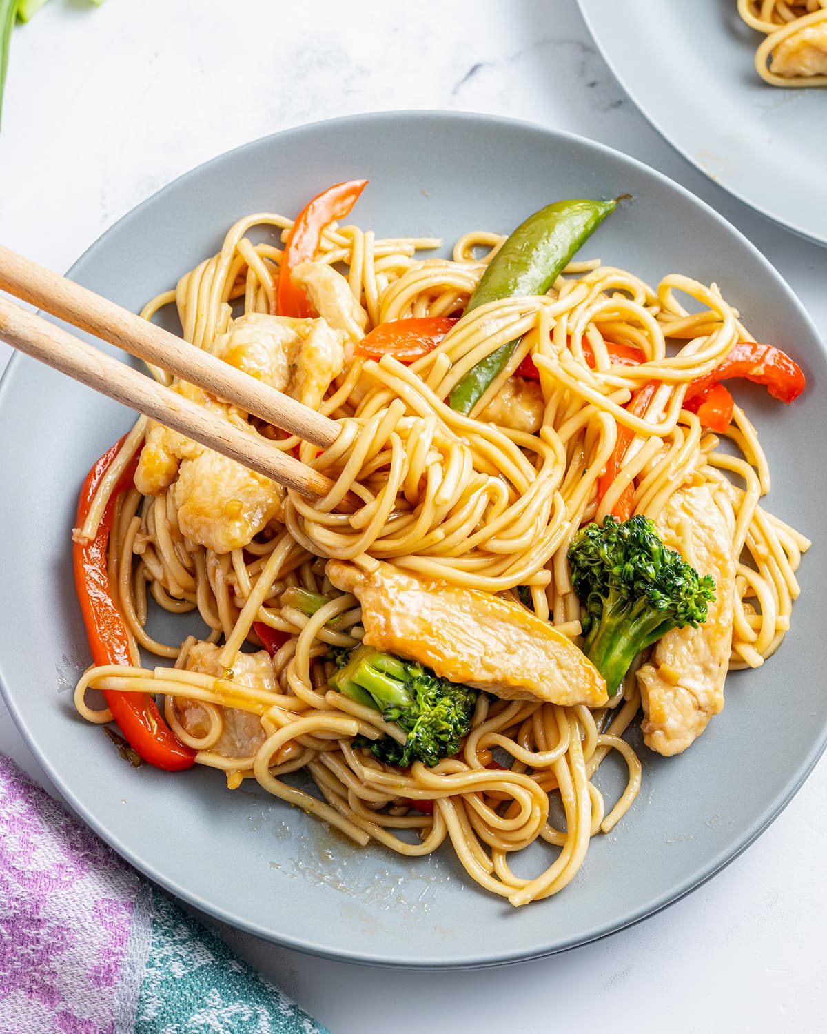 An overhead photo of a plate of chicken lo mein with vegetables with chopsticks wrapping up some of the noodles.