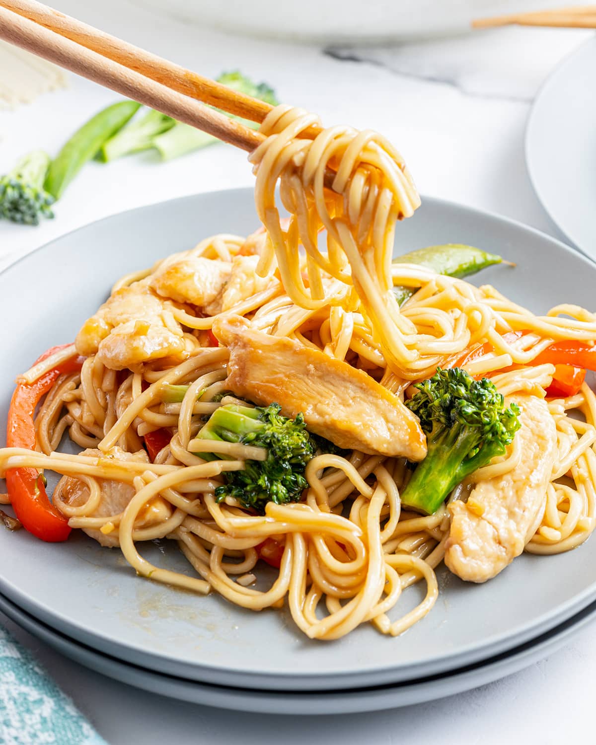A plate of chicken lo mein with broccoli with some of the noodles being lifted out with chopsticks. 
