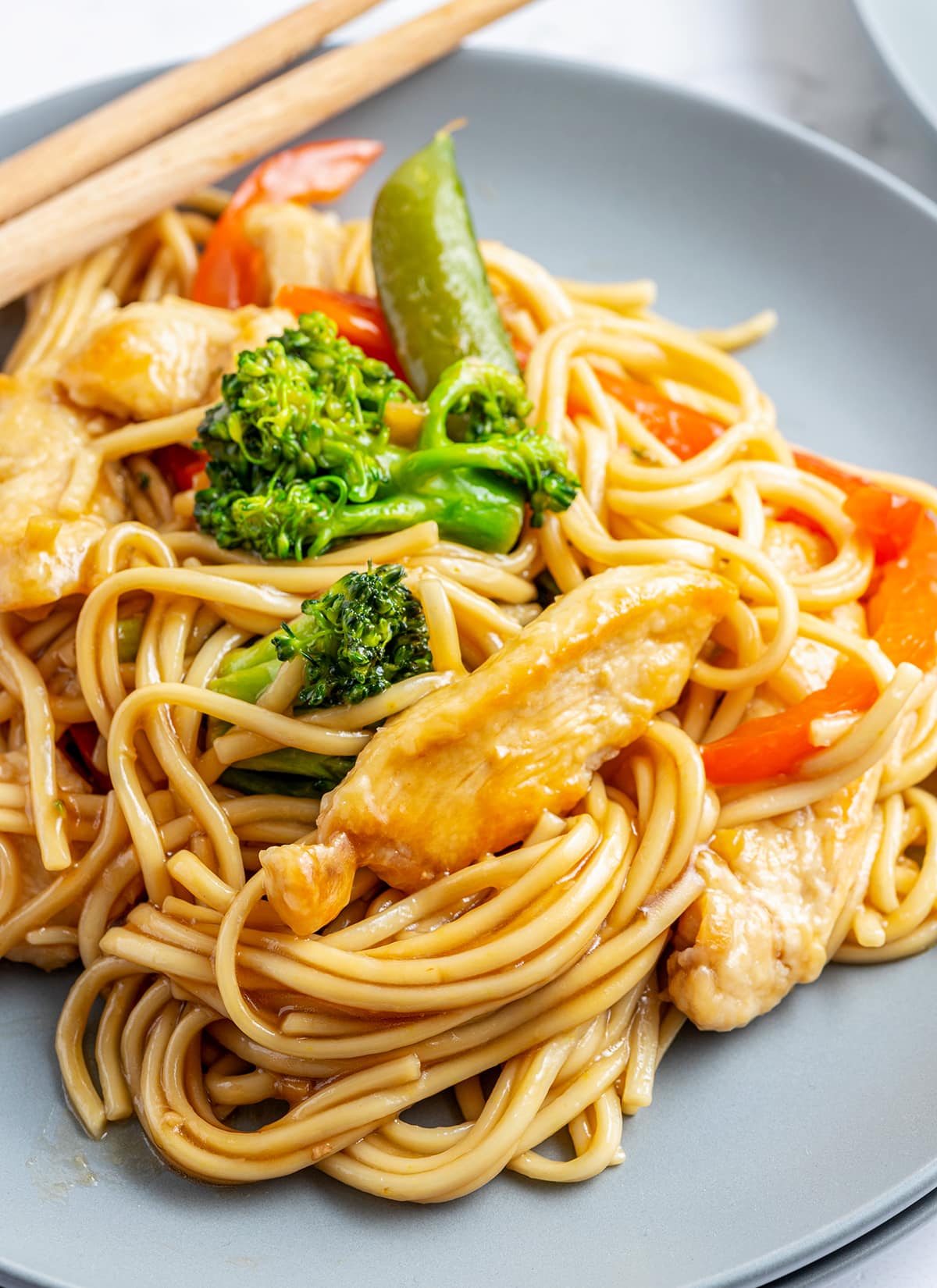A close up of a plate of lo mein with chicken, bell pepper, peas, and broccoli in it. 