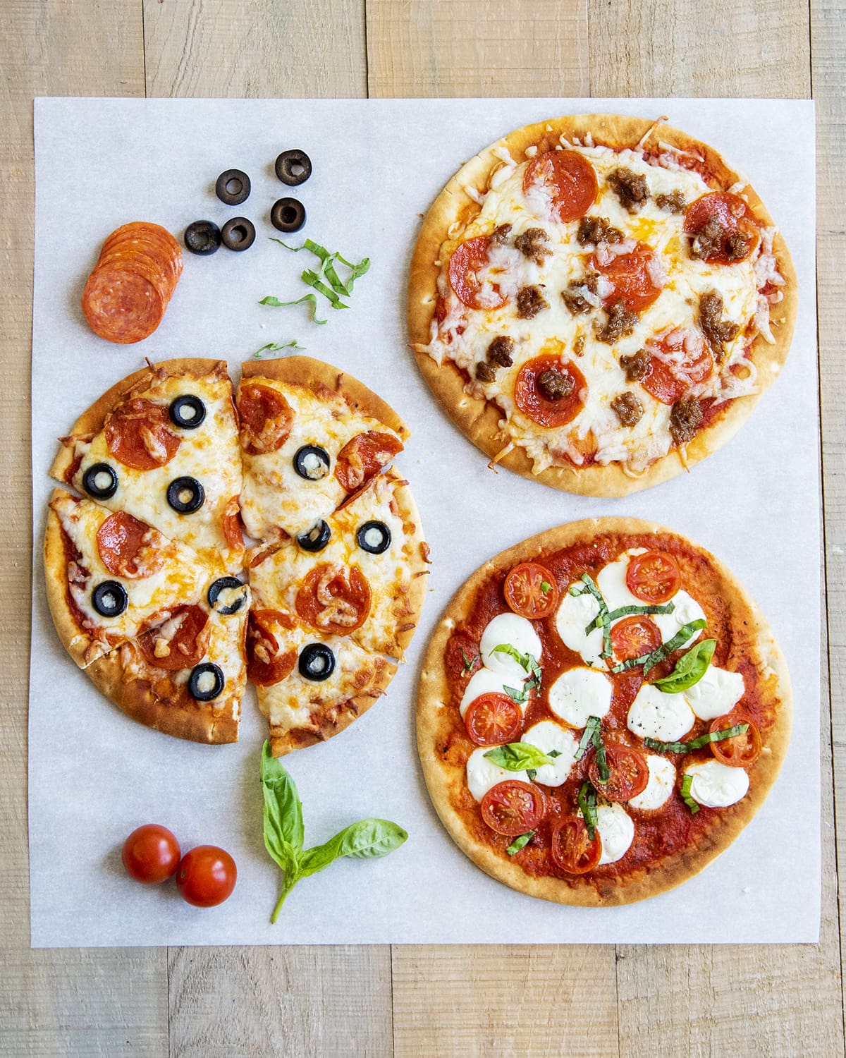 An overhead photo of three pita pizzas topped with pepperoni, sausage, and other pizza toppings on parchment paper.