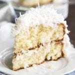 A slice of coconut cake on a white plate. It's a light colored cake, topped with a white frosting, and sweetened coconut.