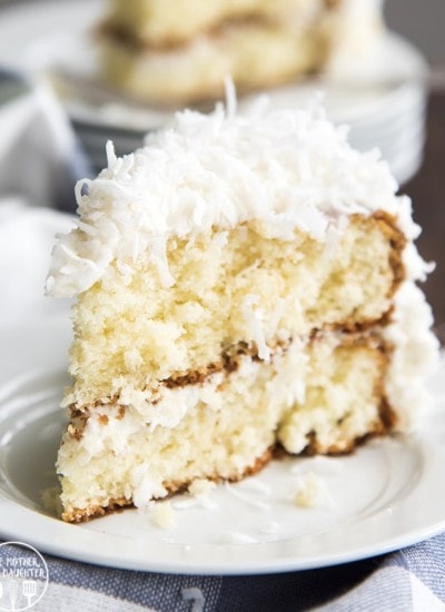 A slice of coconut cake on a white plate. It's a light colored cake, topped with a white frosting, and sweetened coconut.