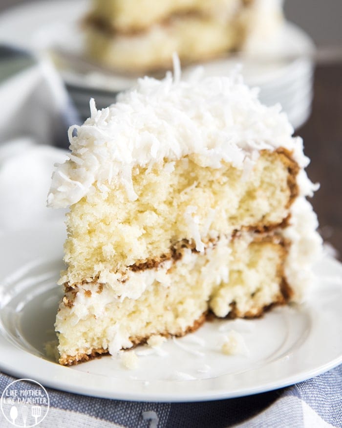 A slice of coconut cake on a white plate. It's a light colored cake, topped with a white frosting, and sweetened coconut.
