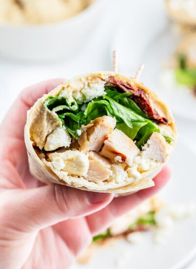 A hand holding half of a chicken wrap, full of lettuce, chicken, sundried tomatoes, and feta.