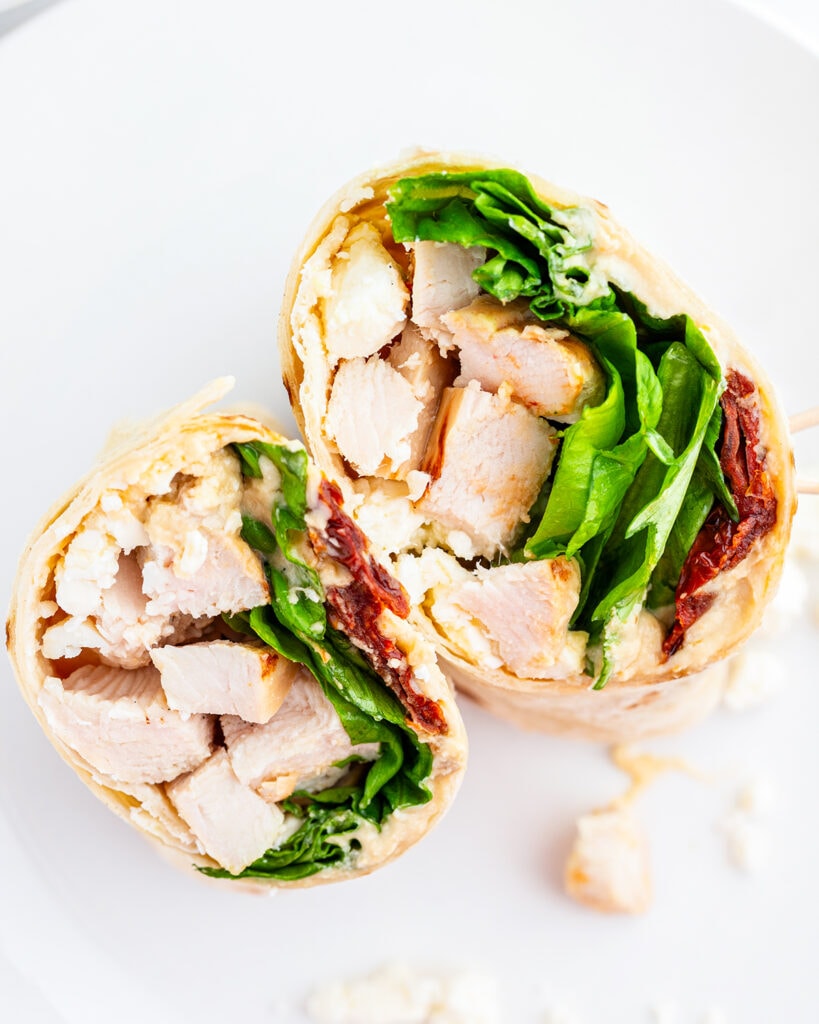 Two halves of a Greek Chicken Wrap with chicken, lettuce cheese, hummus, and sundried tomatoes.