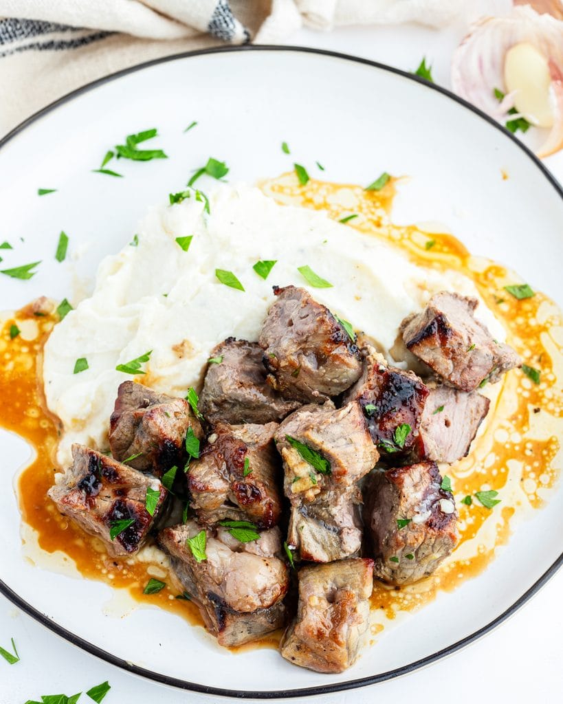 A plate of steak bites on top of a pile of mashed potatoes.