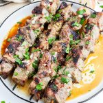 Four steak kabobs on a plate topped with garlic and fresh parsley.