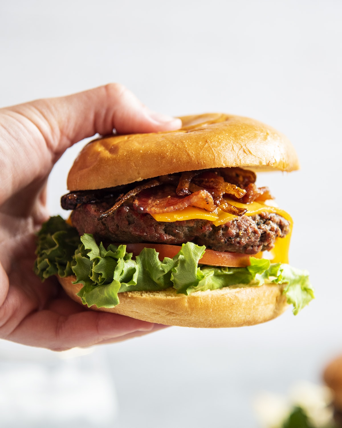 A hand holding a cheeseburger with lettuce, tomato, and topped with bacon and caramelized onions.