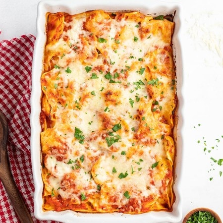 A white baking pan full of pasta with a red sauce, and topped with cheese. There is chopped parsley sprinkled on top.