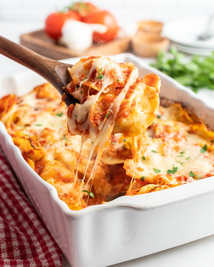 A pan of baked ravioli with a spoon pulling some of the pasta out of the pan, and showing the gooey cheese.