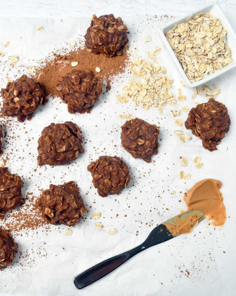 An overview of several no bake cookies on parchment paper with cocoa powder sprinkled about and a smear of peanut butter
