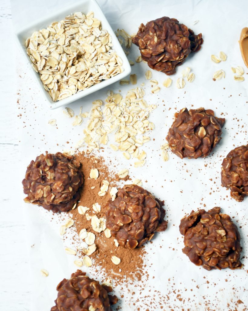 An overview of several no bake cookies on parchment paper with a small bowl of oats to the side and cocoa powder sprinkled about