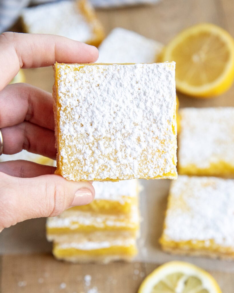 A hand holding a lemon bar square topped with powdered sugar.