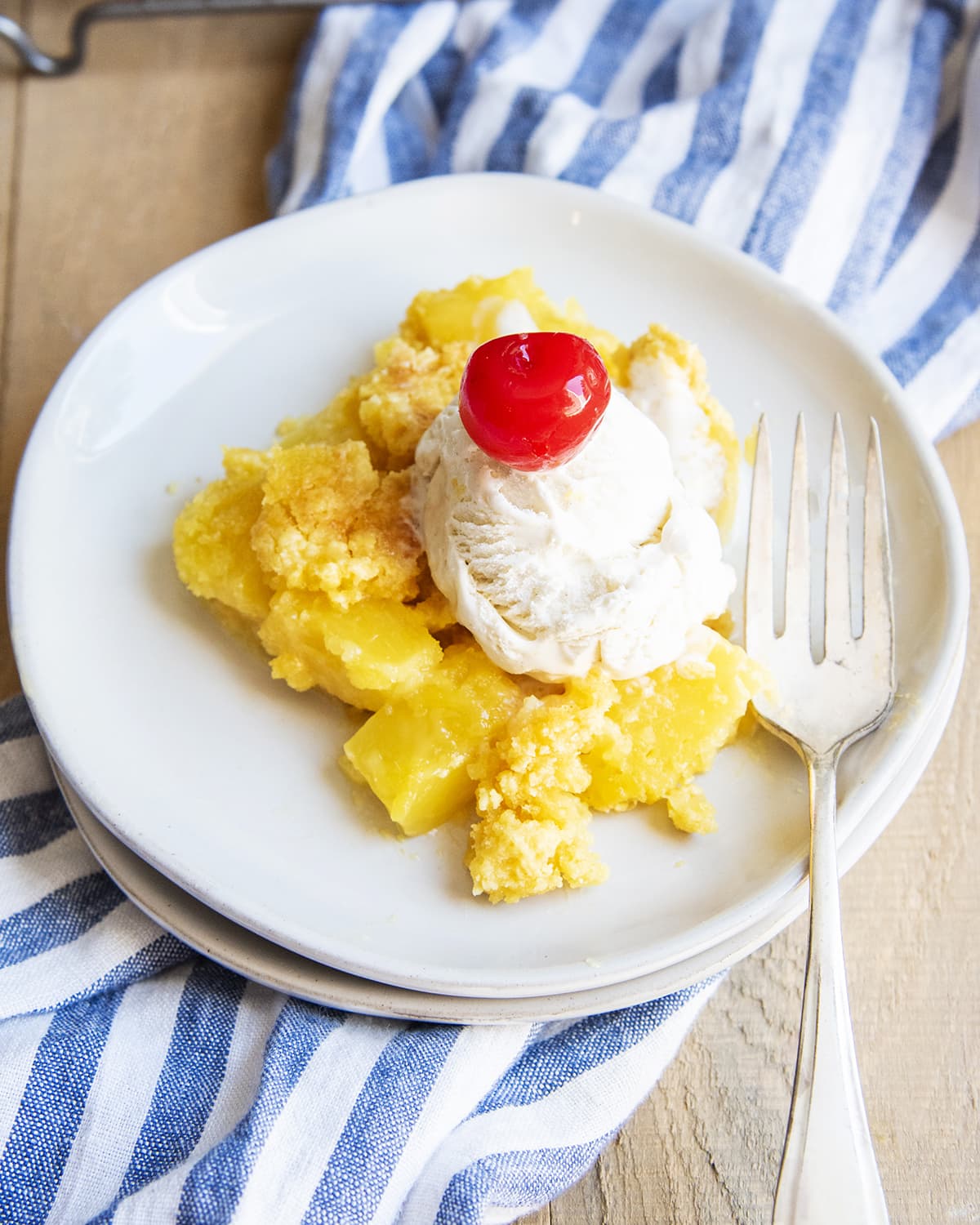 Pineapple dump cake on a plate topped with a scoop of vanilla ice cream and a red cherry.