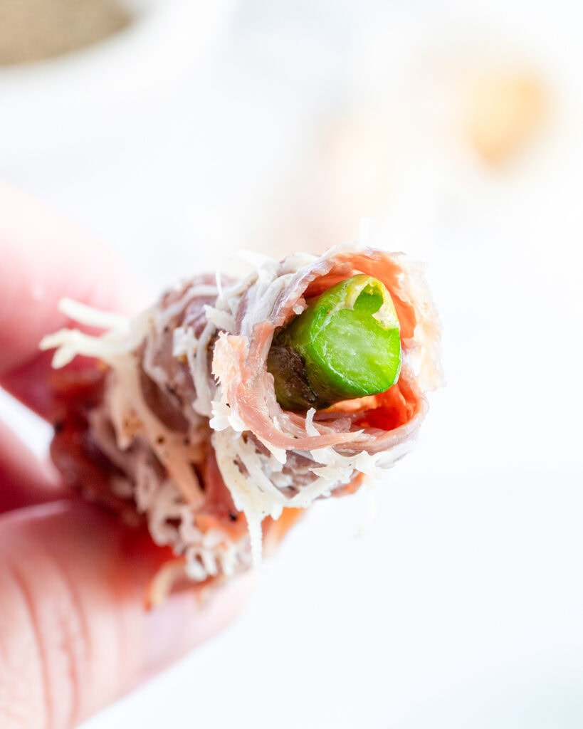 A hand holding an asparagus stalk wrapped in prosciutto.