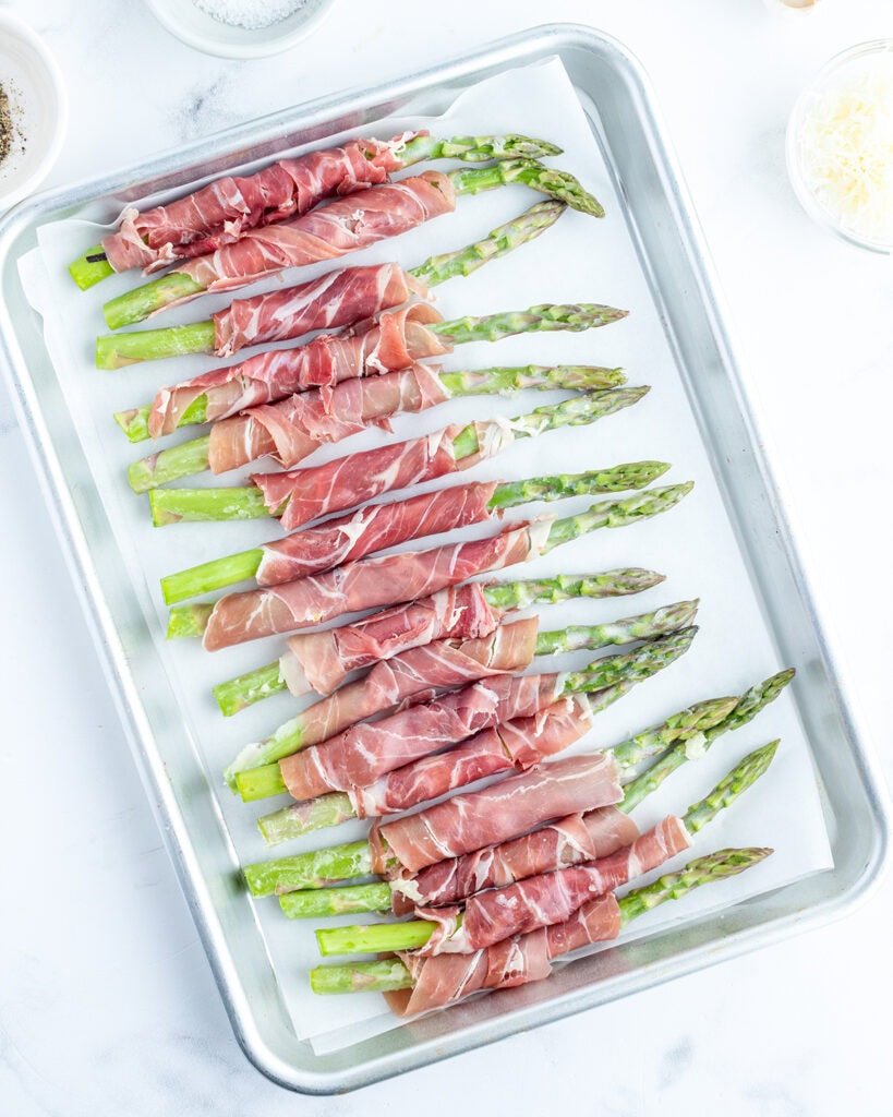 A cookie sheet topped with uncooked green asparagus wrapped in prosciutto.