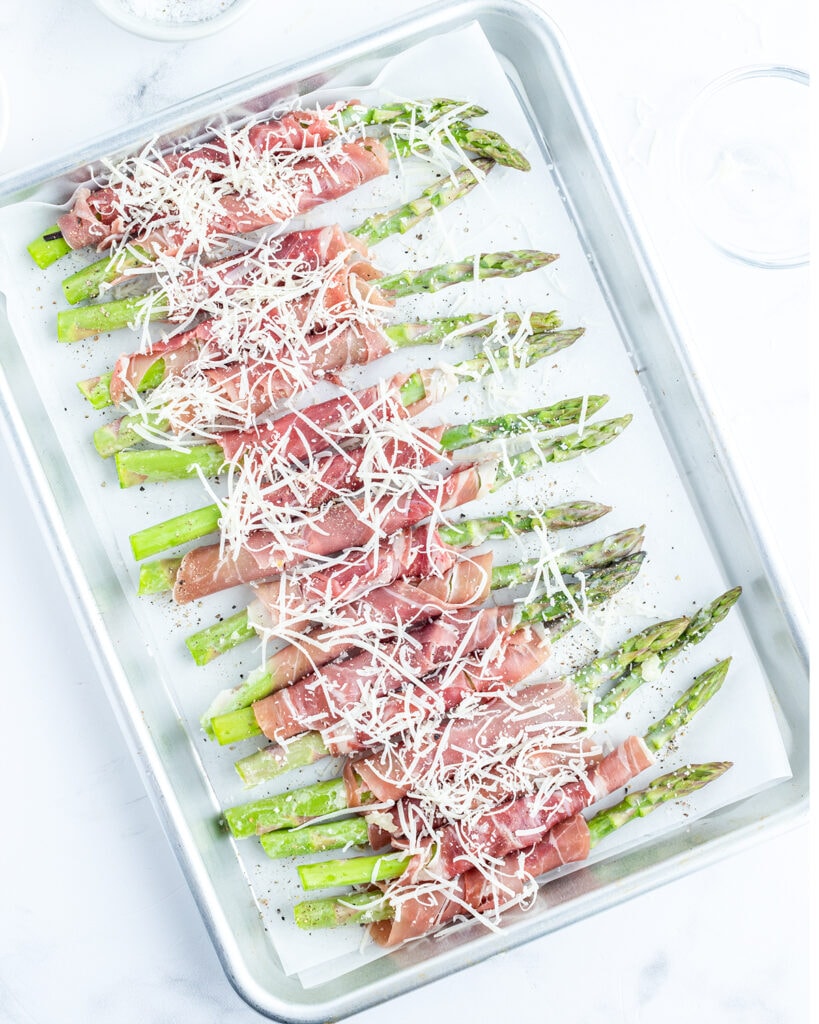 A cookie sheet topped with uncooked green asparagus wrapped in prosciutto and topped with parmesan.