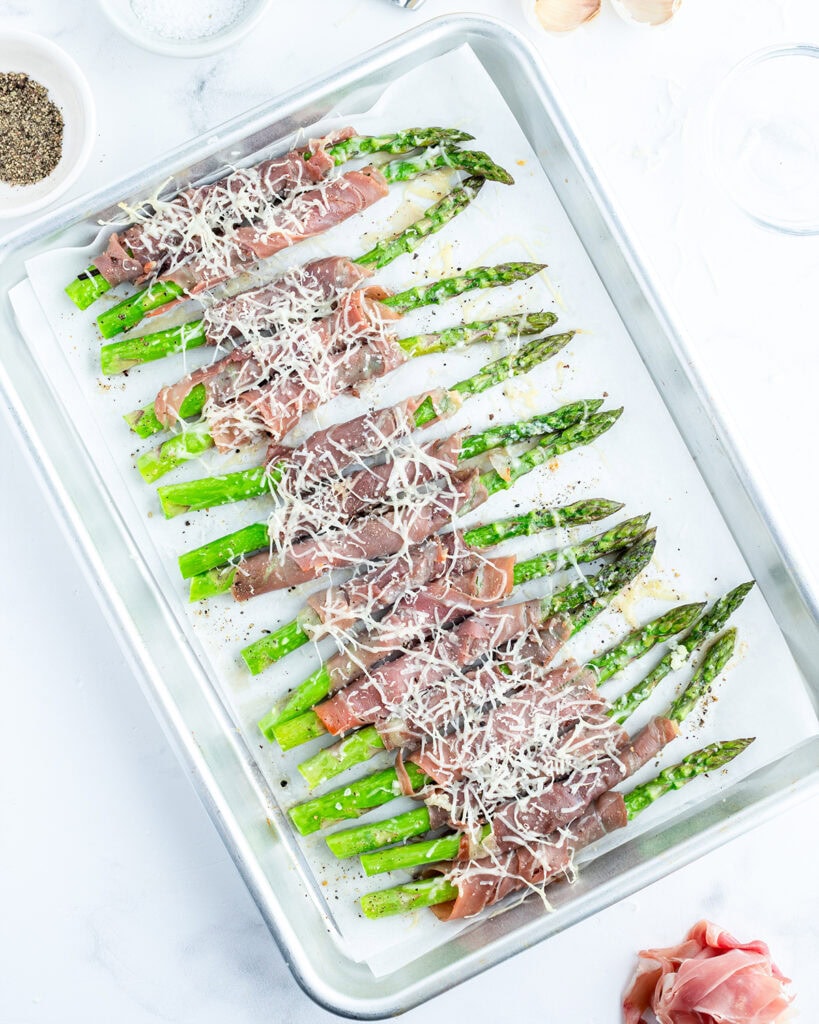 A cookie sheet topped with green asparagus wrapped in prosciutto and topped with parmesan.