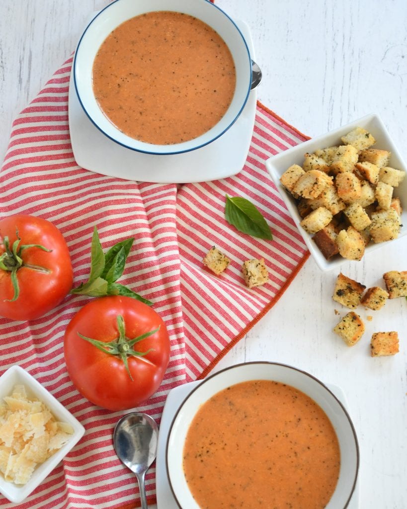 2 bowls of tomato basil so, framed with Parmesan cheese, 2 tomatoes, spoons, bread crumbs on a red and white napkin 