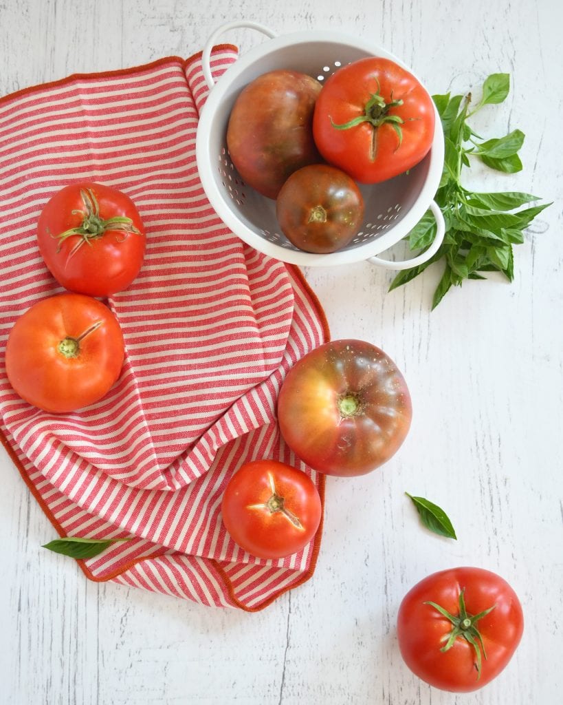 white strainer with 3 tomatoes, 5 large tomatoes, sprigs of fresh basil and basil leaves sprinkled on a red and white napkin