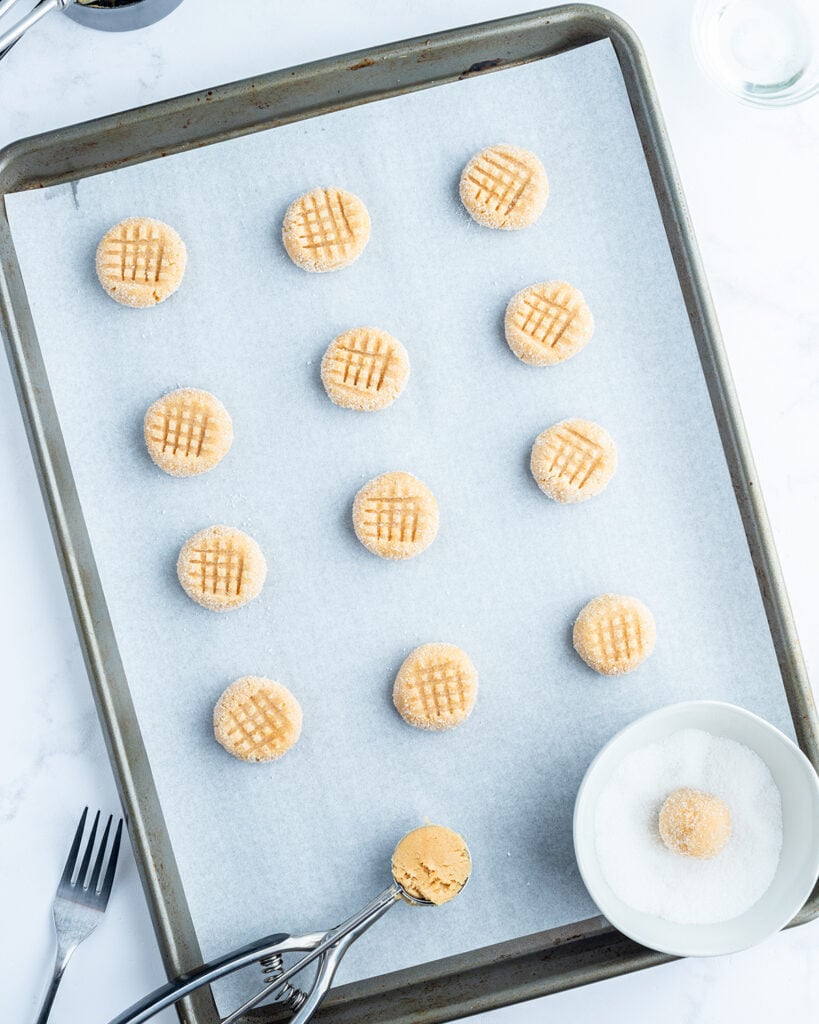 Small peanut butter cookie dough balls with a criss cross pattern on the top on a cookie sheet.