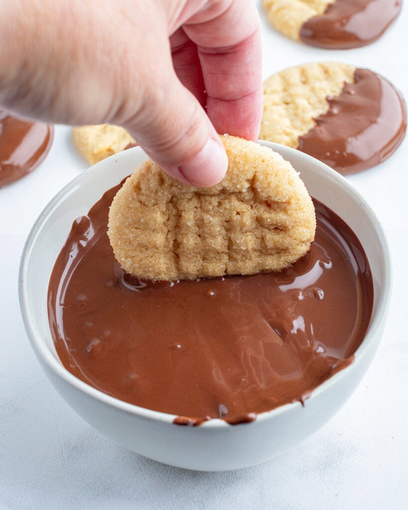A hand holding a peanut butter cookie and dipping it into a bowl of melted chocolate.
