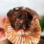 A hand holding a chocolate zucchini muffin with a bite out of it.