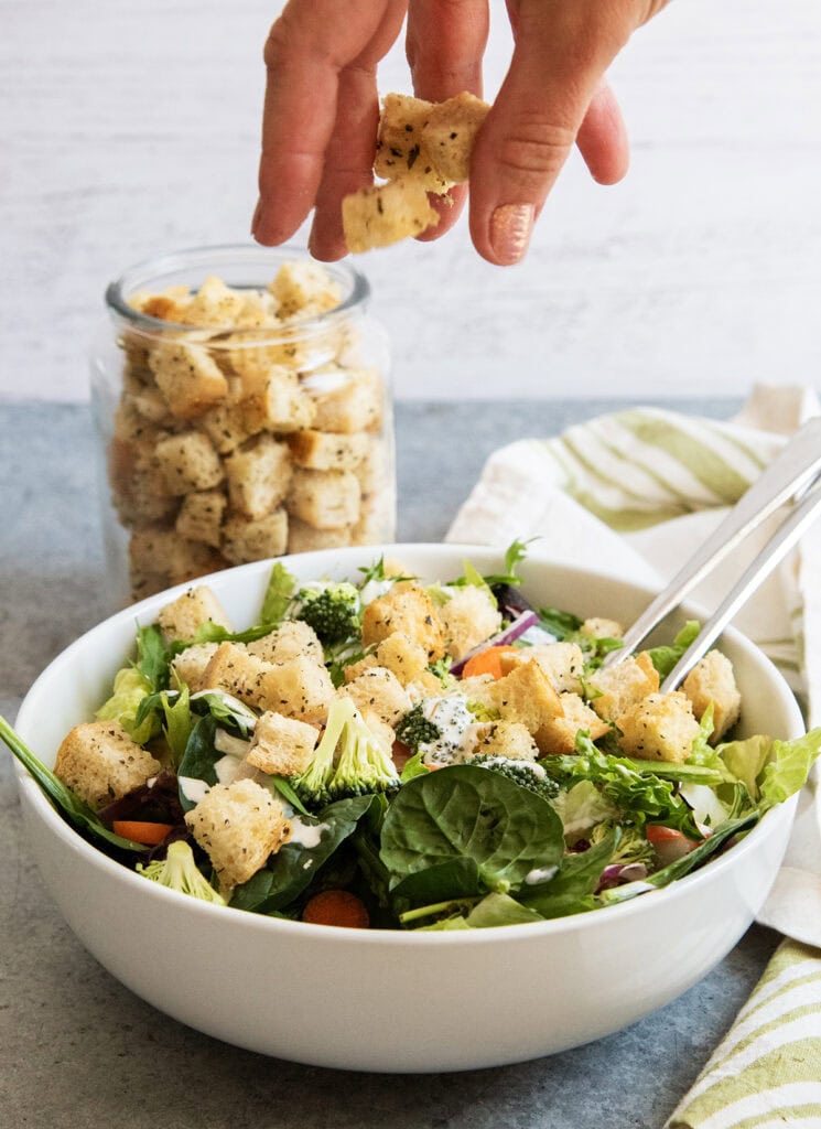 A hand above a bowl of salad sprinkling croutons on top.