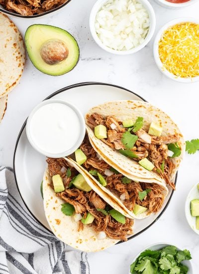 A plate of three pulled pork tacos topped with avocado and cilantro.