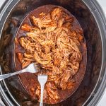 An overhead photo of a slow cooker full of pulled pork in a seasoned tomato sauce.