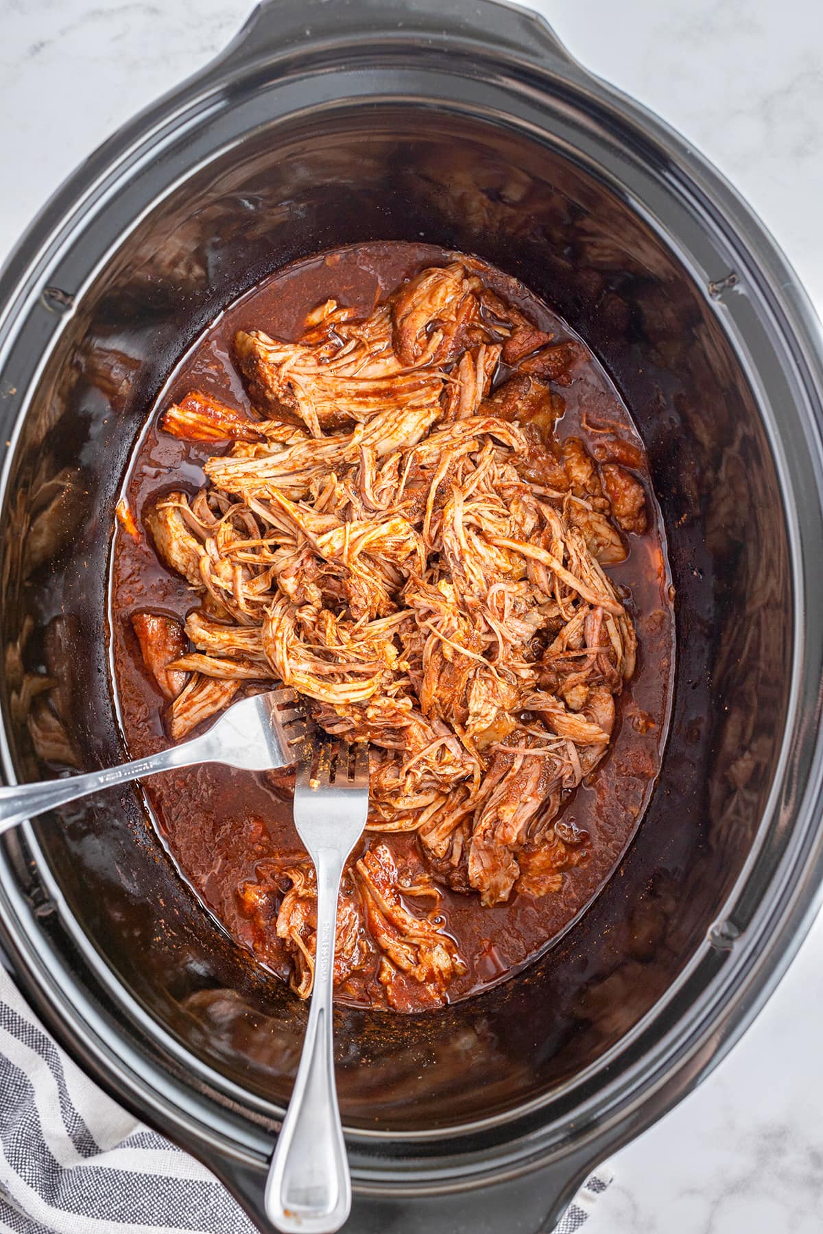 An overhead photo of a slow cooker full of pulled pork in a seasoned tomato sauce.