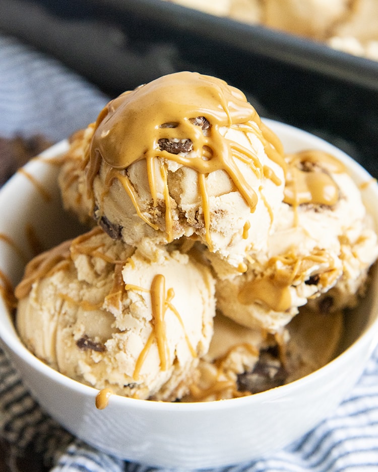 A bowl of peanut butter ice cream, with peanut butter swirls, and peanut butter cups, drizzled with more peanut butter over the top.