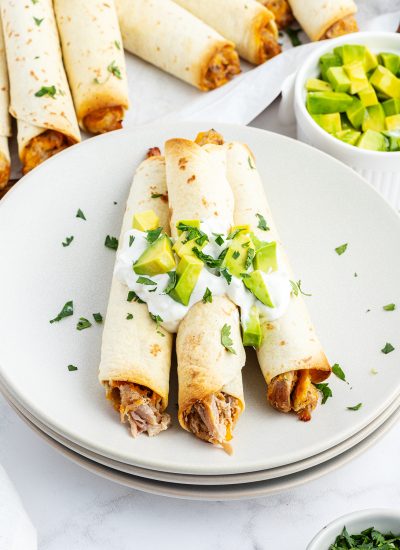 A plate of creamy pulled pork taquitos topped with sour cream and avocado pieces.