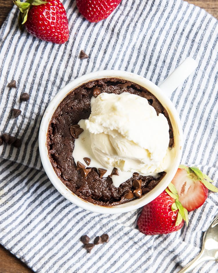An overhead shot of a microwave brownie in a mug with vanilla ice cream with strawberries on the side